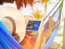 A woman in a hammock in a tropical destination is looking at an iPad showing Time Together, the Grand Pacific Resorts owner magazine.
