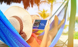 A woman in a hammock in a tropical destination is looking at an iPad showing Time Together, the Grand Pacific Resorts owner magazine.