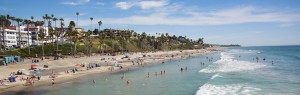 A shot from the San Clemente Pier shows the water full of swimmers, a beautiful blue sky, and the coastal bluffs.