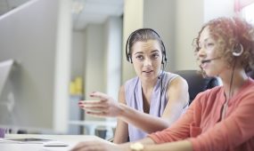 Two women sit together at a desk while one shows the other how to serve as a call center agent.