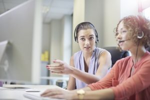 Two women sit together at a desk while one shows the other how to serve as a call center agent.